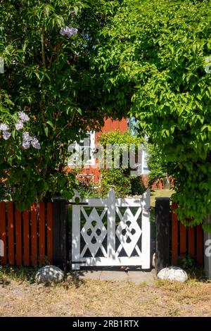 White wooden gate on falu red paint fence leading to a garden below lush green trees in hot Swedish summer in rural countryside setting Stock Photo