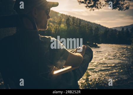 Back View of Relaxed Man Sitting in Camping Chair by the River, Enjoying Sunshine and Beautiful Wildlife Scenery. Stock Photo