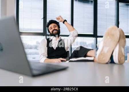 Portrait of happy Indian businessman sitting at office desk with feet up on table with laptop, finished work, job done, completed all tasks Stock Photo