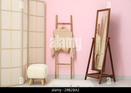 Sweater hanging on wooden ladder, folding screen, bench and mirror near pink wall in room Stock Photo