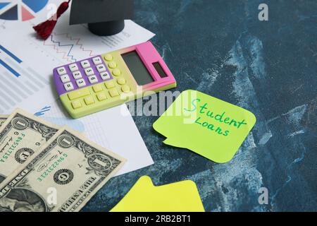 Sticky note with text STUDENT LOANS, calculator, dollar banknotes and graduation cap on blue table Stock Photo