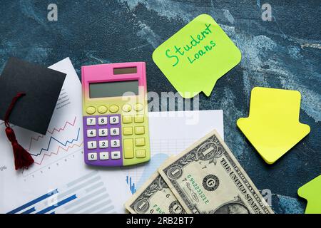 Sticky note with text STUDENT LOANS, calculator, dollar banknotes and graduation cap on blue table Stock Photo