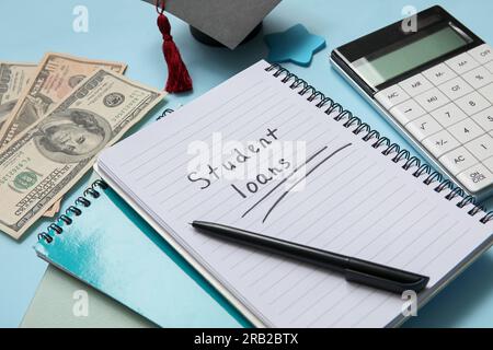Notebook with text STUDENT LOANS,  calculator, dollar banknotes and graduation cap on blue background Stock Photo