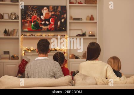 Family watching Christmas movie via TV in cosy room, back view. Winter holidays atmosphere Stock Photo