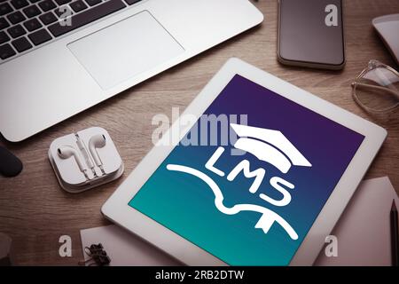 Learning management system. Tablet with open book and graduate cap icons with abbreviation LMS on screen. Workplace with different devices on wooden t Stock Photo