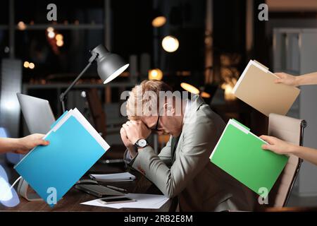 Deadline concept. Colleagues giving folders with documents to overwhelmed man at desk in evening Stock Photo