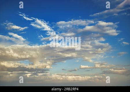Blue sky with wispy white cumulus clouds scattered across the horizon illuminated by the sun, which is setting in the distance, casting a soft orange Stock Photo