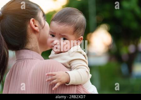 mother holding and comforting her crying infant baby outside in the park with sunlight Stock Photo