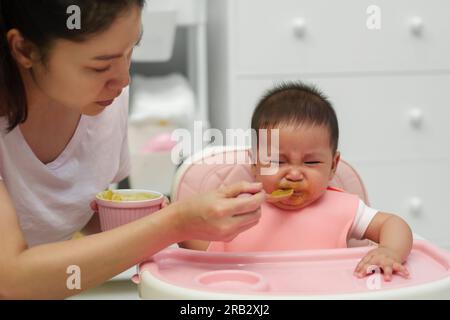 mother feeding food to her crying infant baby with a spoon, refuse to eat Stock Photo