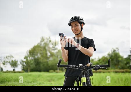A happy Asian man in sportswear and a bike helmet is on his bike and using an online map on his smartphone. Stock Photo