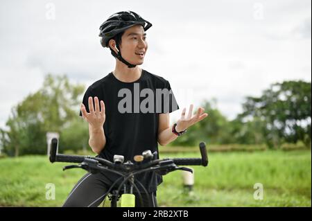 A handsome Asian man in sportswear and earbuds is talking with someone or dealing business on the phone while riding a bike in the nature park on the Stock Photo
