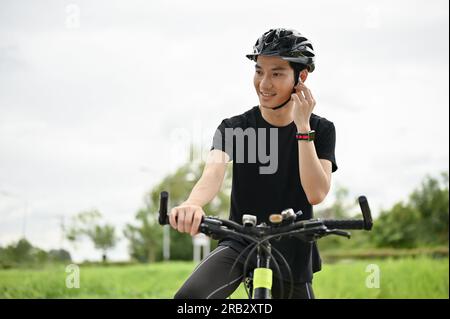 A happy and handsome young Asian man wearing his earbuds, listening to music while riding a bike in the nature park. Stock Photo