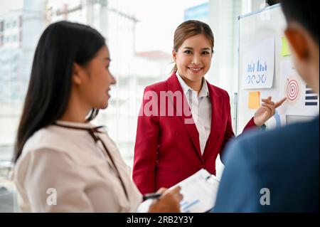 A confident millennial Asian businesswoman presented her marketing project on a whiteboard to her team in the meeting. creative brainstorming meeting Stock Photo