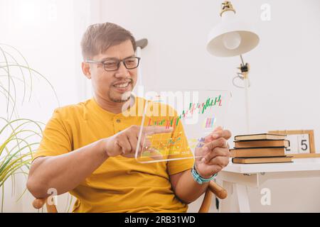 Happy southeast asian business male looking at financial stock chart investment casual holiday at home Stock Photo