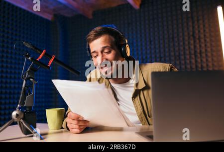 Smiling radio host with headphones reading news from paper into studio microphone at radio station with neon lights. looking at camera Stock Photo