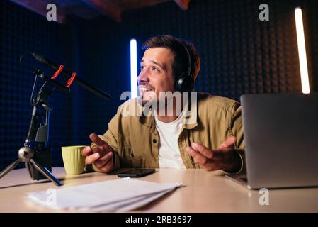 Young stylish man in mustard shirt with headphones gesturing at microphone and sharing story with audience while sitting at desk in studio with neon l Stock Photo