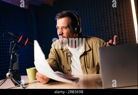 Gesturing radio host with headphones reading news from paper into studio microphone at radio station with neon lights Stock Photo