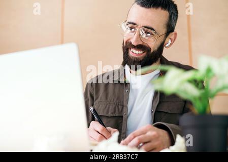 Concentrated man in glasses taking notes writing information from laptop in cafe, cheerful bearded man studying in cafe Stock Photo