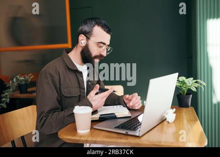 Stylish bearded man sitting at table with laptop and cup of coffee while spending time in modern cafeteria and working on distance Stock Photo