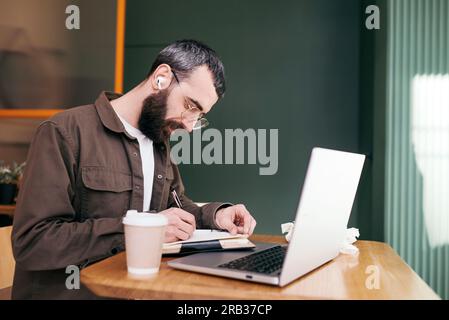 Stylish bearded man sitting at table with laptop and cup of coffee while spending time in modern cafeteria and working on distance Stock Photo