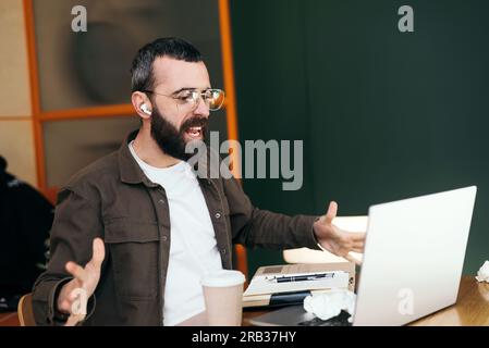 Angry caucasian male freelancer in a cafe quarreling while talking on webcam, discussing problems with an online business project Stock Photo
