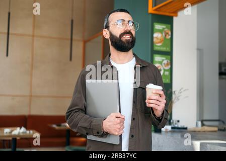 Smiling bearded creative man in glasses standing holding laptop and cup of coffcee in cafe looking out window. Freelancer and modern interior Stock Photo