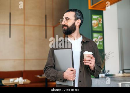 Smiling bearded creative man in glasses standing holding laptop and cup of coffee in cafe. Freelancer and modern interior Stock Photo