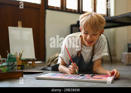 Carefree teenage male lying on floor in art studio and painting on canvas with watercolors. Creative hobby and art concept Stock Photo