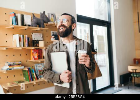 Stylish bearded man in glasses standing holding laptop and cup of coffee in cafe and smiling. Freelancer Stock Photo