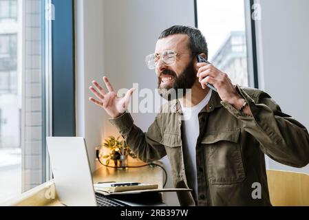 Angry caucasian male freelancer in cafe having quarrel during mobile phone conversation discussing problems with online business project Stock Photo