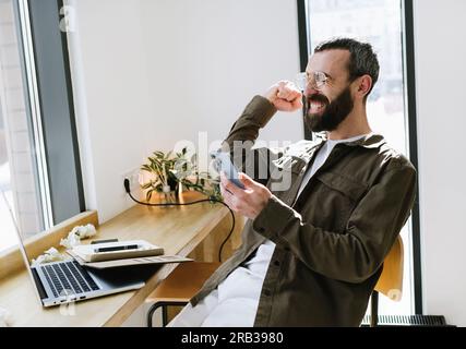 Cheerful bearded man in glasses rejoices in a cafe using a mobile phone, making a winner gesture. Distant work. Coffee shop, remote work, freelance Stock Photo