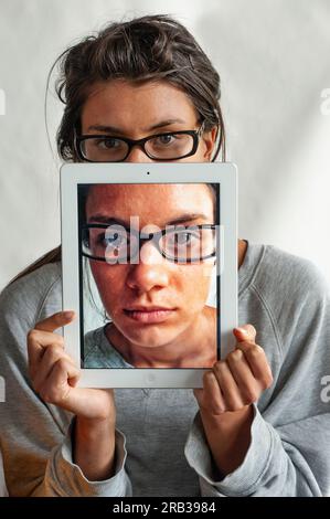 A young woman using her iPadto hide behind,  on which there is a photo of her face Stock Photo