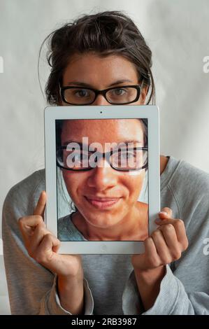 A young woman using her iPadto hide behind,  on which there is a photo of her face Stock Photo