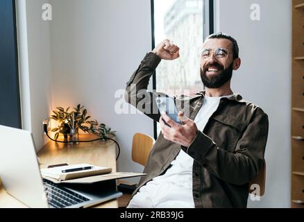 Cheerful bearded man in glasses rejoices in a cafe using a mobile phone, making a winner gesture. Distant work. Coffee shop, remote work, freelance Stock Photo