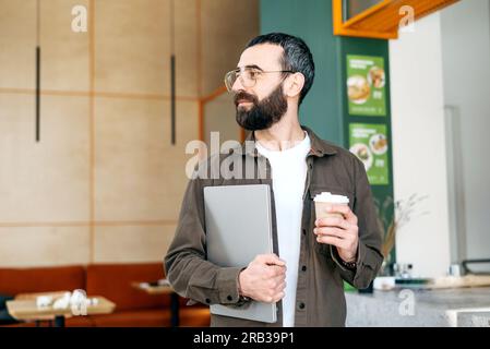 Smiling bearded man in glasses standing holding laptop and cup of coffee in cafe looking out window. Freelancer Stock Photo