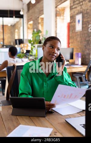 Focused african american casual businesswoman talking on smartphone with document in creative office. Business, work and technology, unaltered. Stock Photo