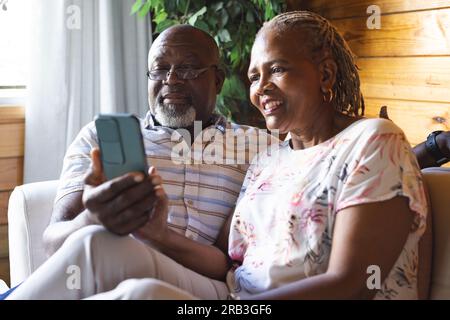 Happy senior african american couple sitting on sofa and using smartphone at home. Senior lifestyle, relaxing, communication and retirement, unaltered Stock Photo