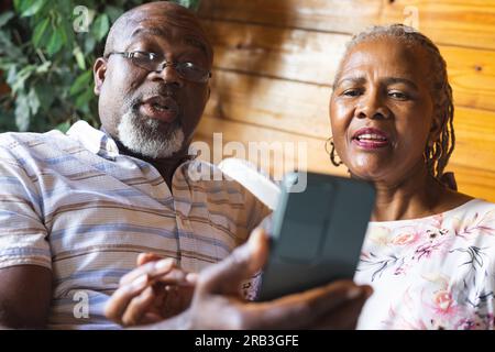 Senior african american couple sitting on sofa and using smartphone at home. Senior lifestyle, relaxing, communication and retirement, unaltered. Stock Photo