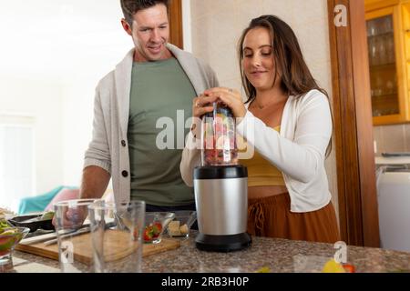 Happy caucasian couple mixing fruits using blender in kitchen. Healthy lifestyle, food and domestic life, unaltered. Stock Photo
