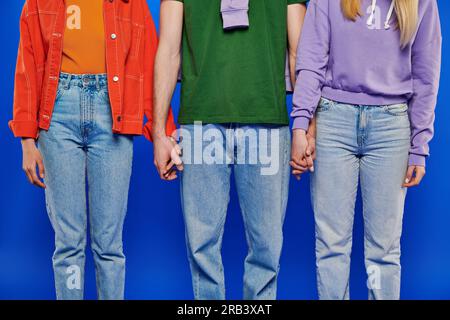 alternative family, cropped view of three polygamy people, young man and women holding hands on blue background, studio shot, vibrant clothes, polyamo Stock Photo