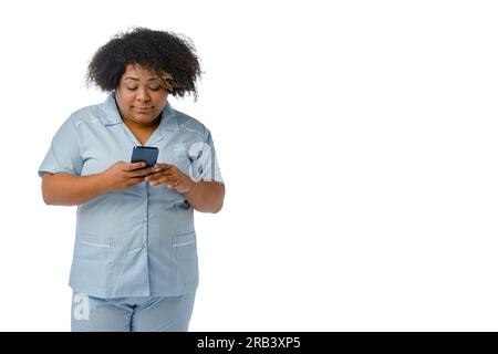 young Afro-Latin female doctor in blue uniform, is standing watching medical appointments on the phone, reading messages and notifications, white back Stock Photo