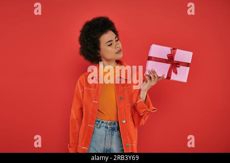 holiday, confused african american woman in casual attire holding wrapped present on coral background, vibrant colors, gift box, attractive and stylis Stock Photo