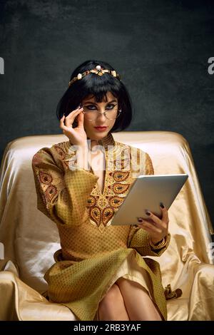 Portrait of young beautiful woman, queen, Cleopatra in golden dress sitting  with tablet against dark vintage background. Business lady Stock Photo -  Alamy
