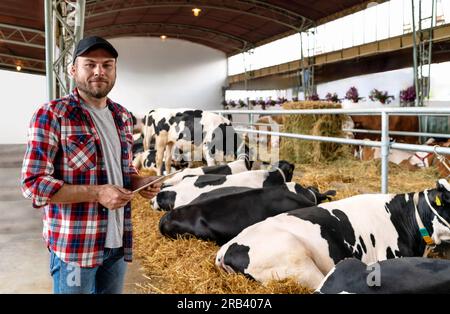 Portrait young adult farmer holding digital tablet in hands and smiling against background of cows in paddock in livestock farm. Stock Photo