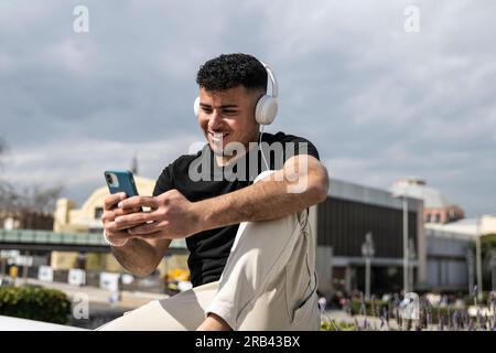Young handsome man sitting on bank relaxed and texting with her phone using headphones. Satisfied guy smiling and using an smartphone sitting in the Stock Photo