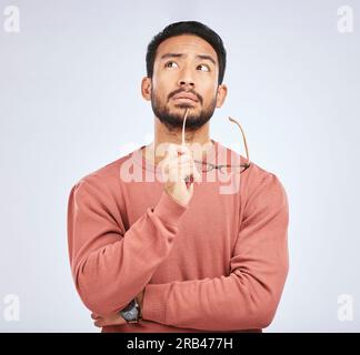 Decision, thinking and asian man in studio with glasses, questions and brainstorming on grey background. Why, idea and male person with emoji, body Stock Photo