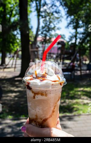 Woman hand holding a transparent plastic cup with a caramel frappe on a defocused park background Stock Photo