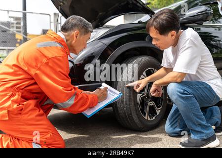 A man pointing out the damage on his car's tyre from the car accident to the car insurance mechanic wearing yellow hardhat and orange jumpsuit or agen Stock Photo