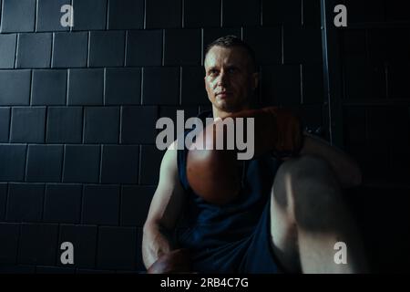 Tired boxer in boxing gloves resting in dark gym after exercising. Stock Photo