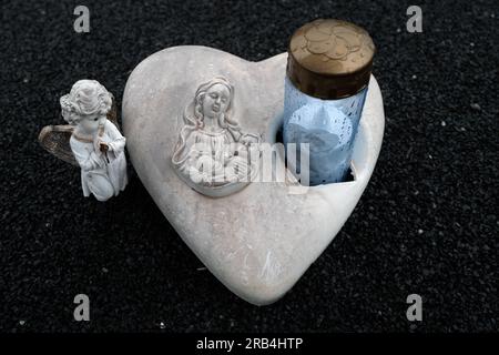 A heart-shaped stone with the image of the Blessed Virgin Mary and Baby Jesus, flanked by two small statues of angels on a black sand background Stock Photo
